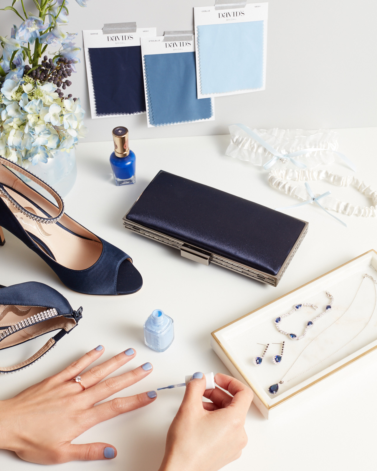 Hand being painted with light blue nail polish with blue heels, handbag, and jewelry on table.