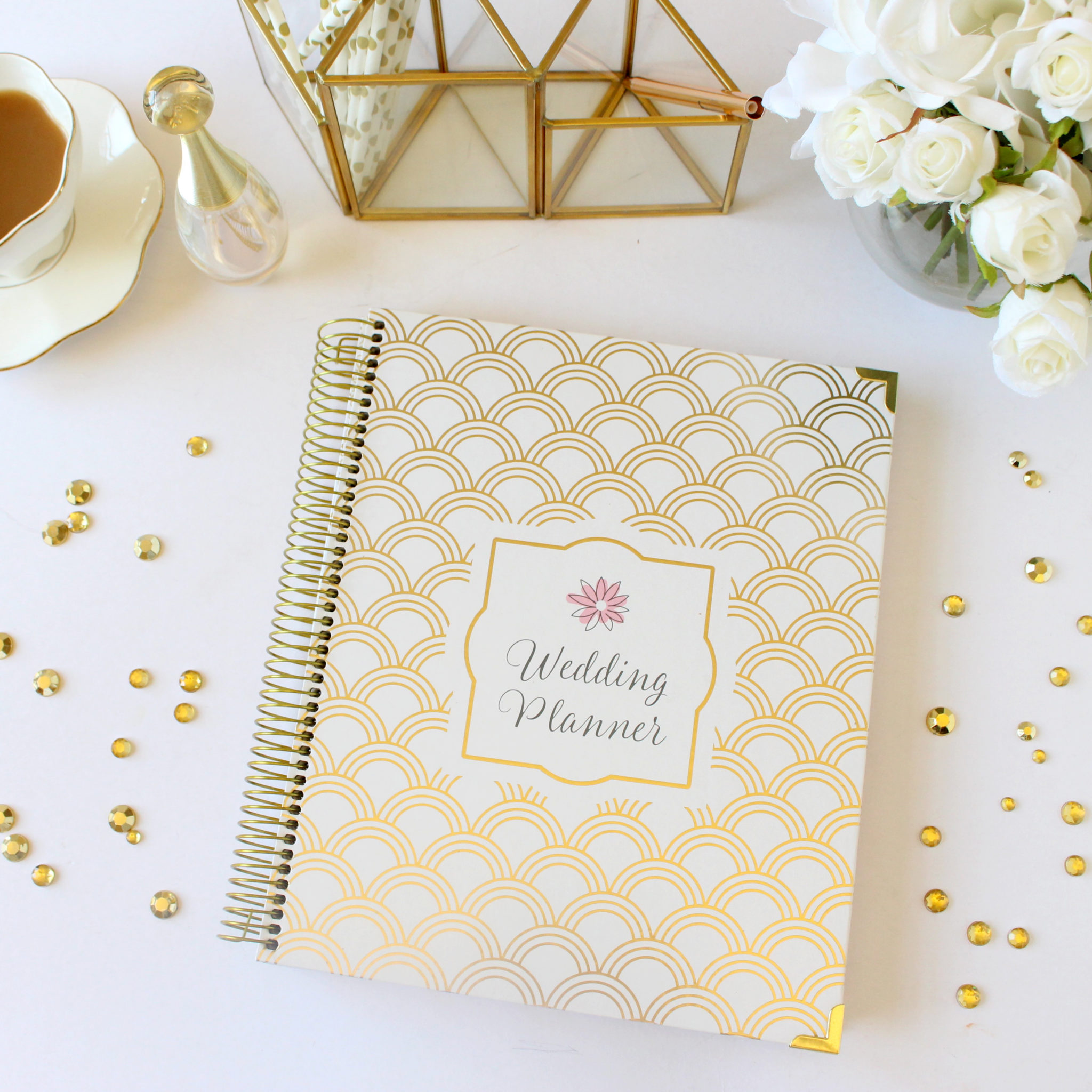 White and gold wedding planning notebook on a white table with a white and gold tea cup and vase of white roses