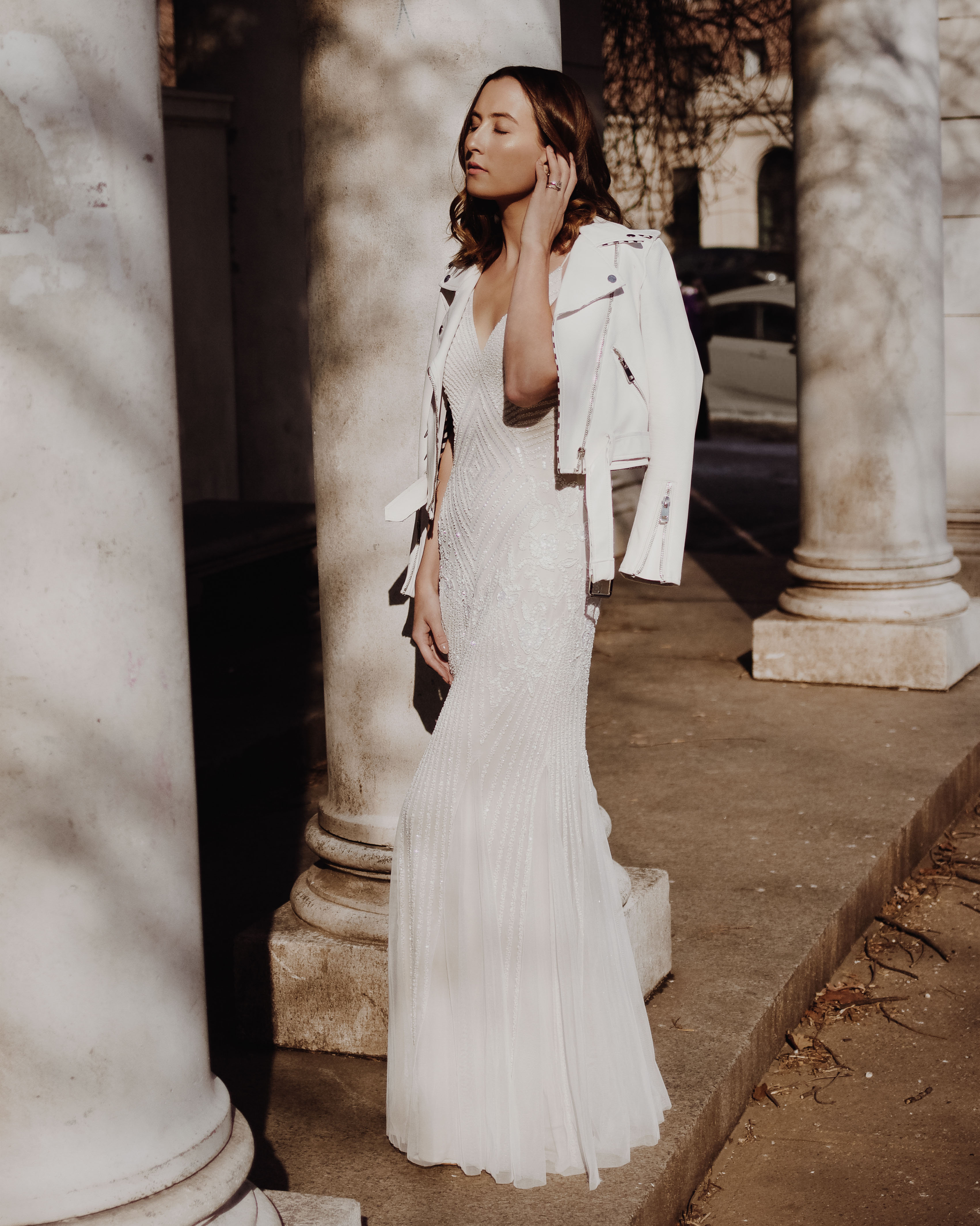 Woman standing near white pillars in a long white beaded dress with white leather jacket draped over her shoulders