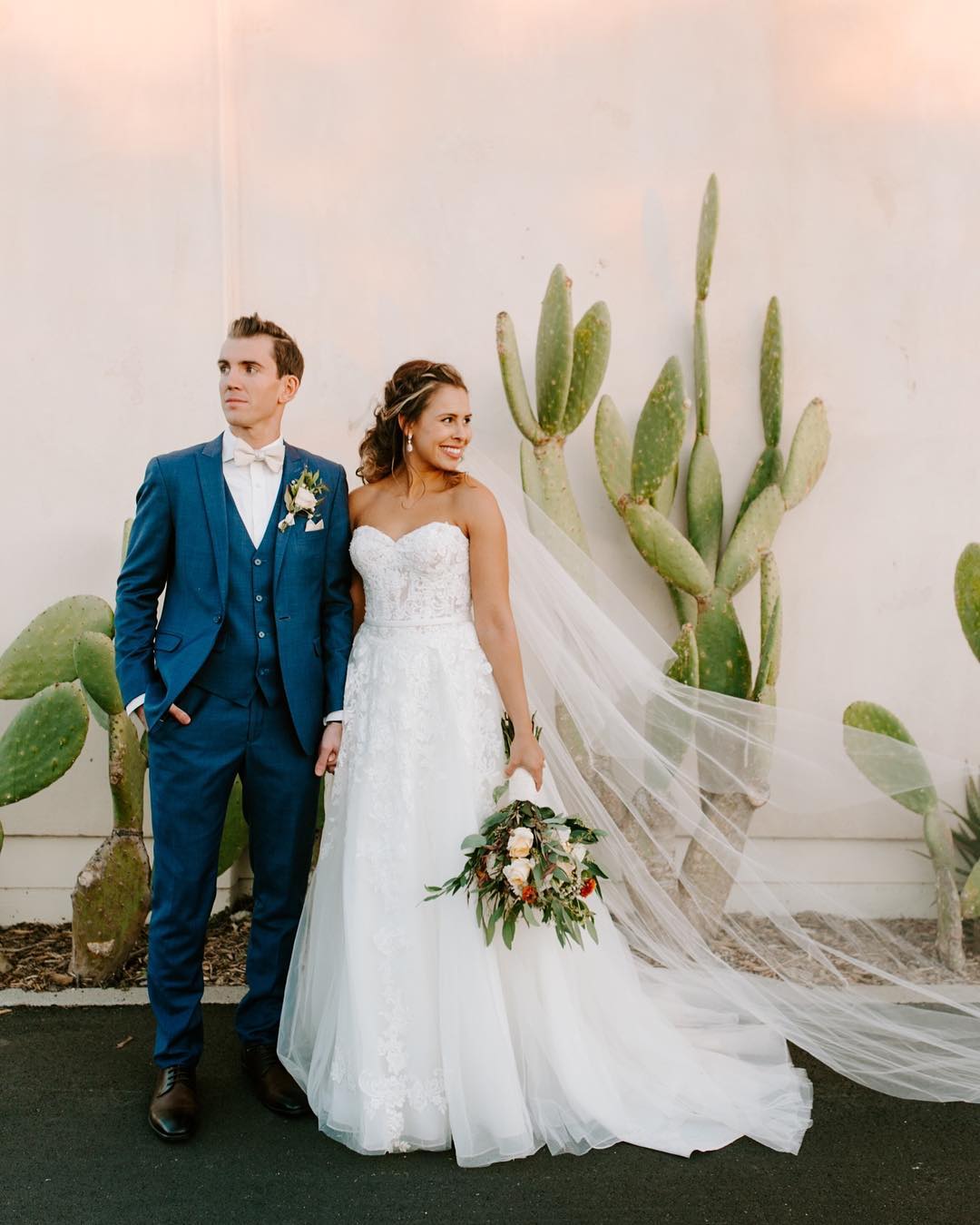 Bride in strapless ballgown with groom in blue three-piece suit stand near a cactus. 