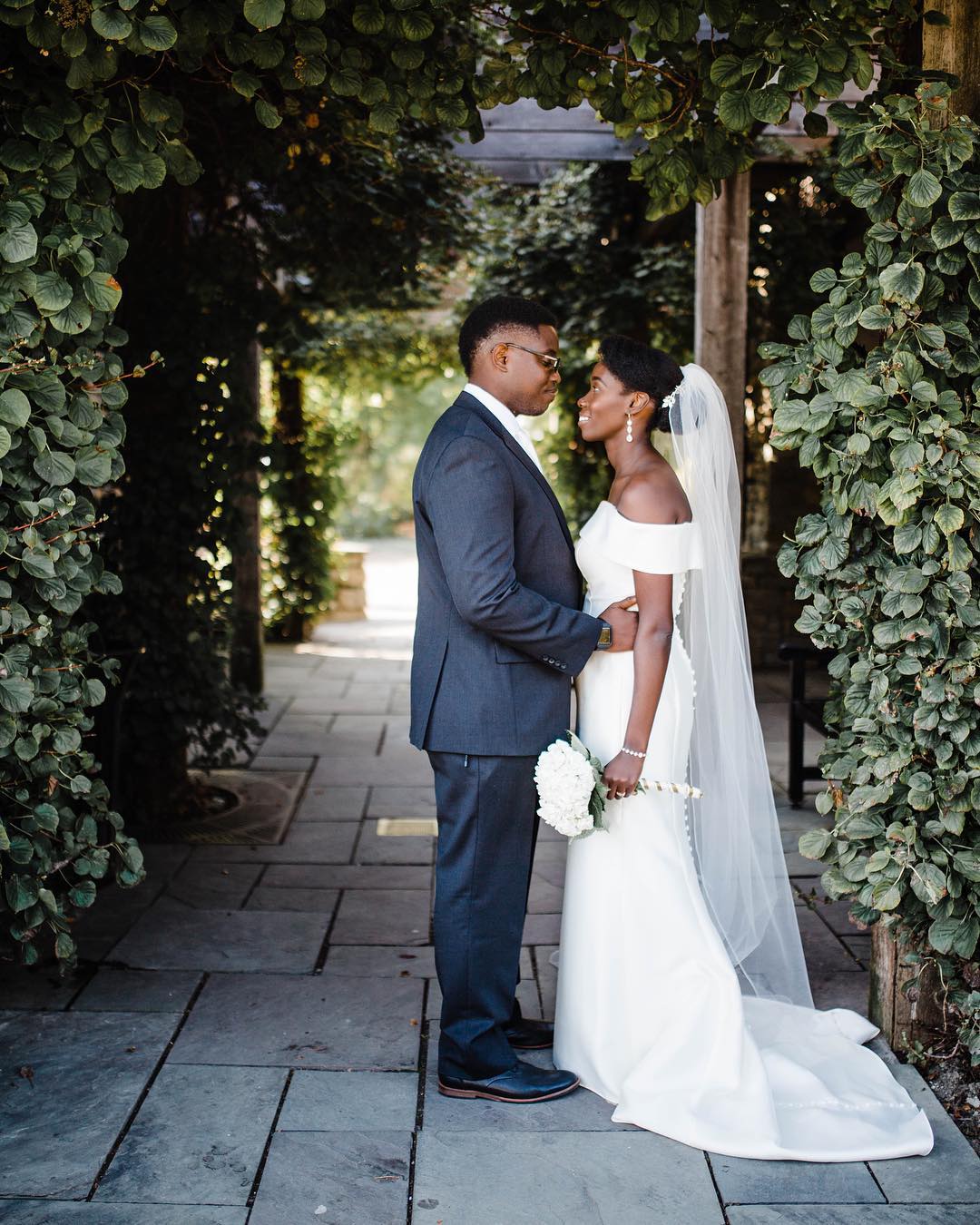 African American bride and groom standing under an archway covered in greenery. 