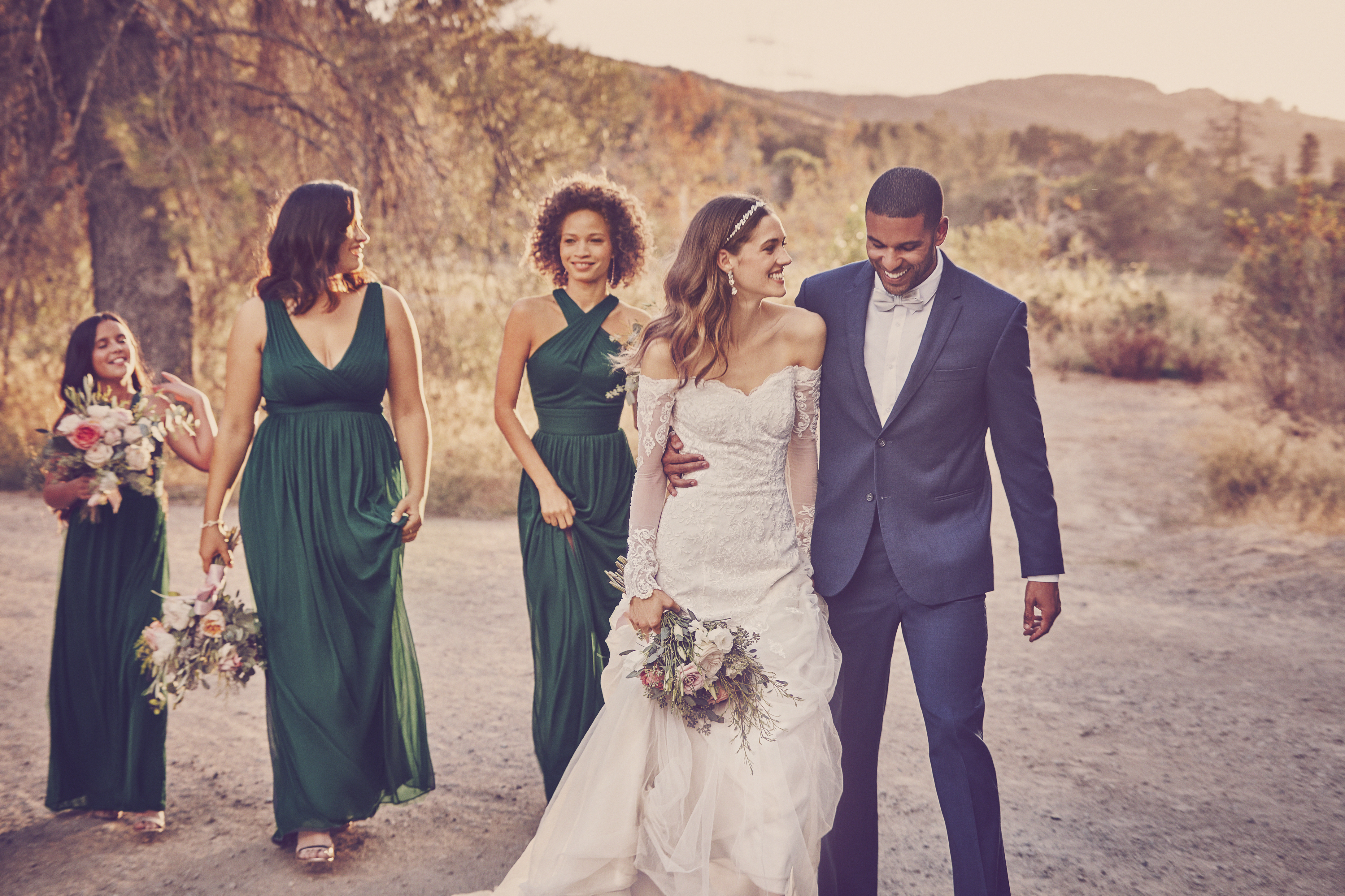 Man in suit, bride in white dress, and bridesmaids in green dresses walking on dirt road