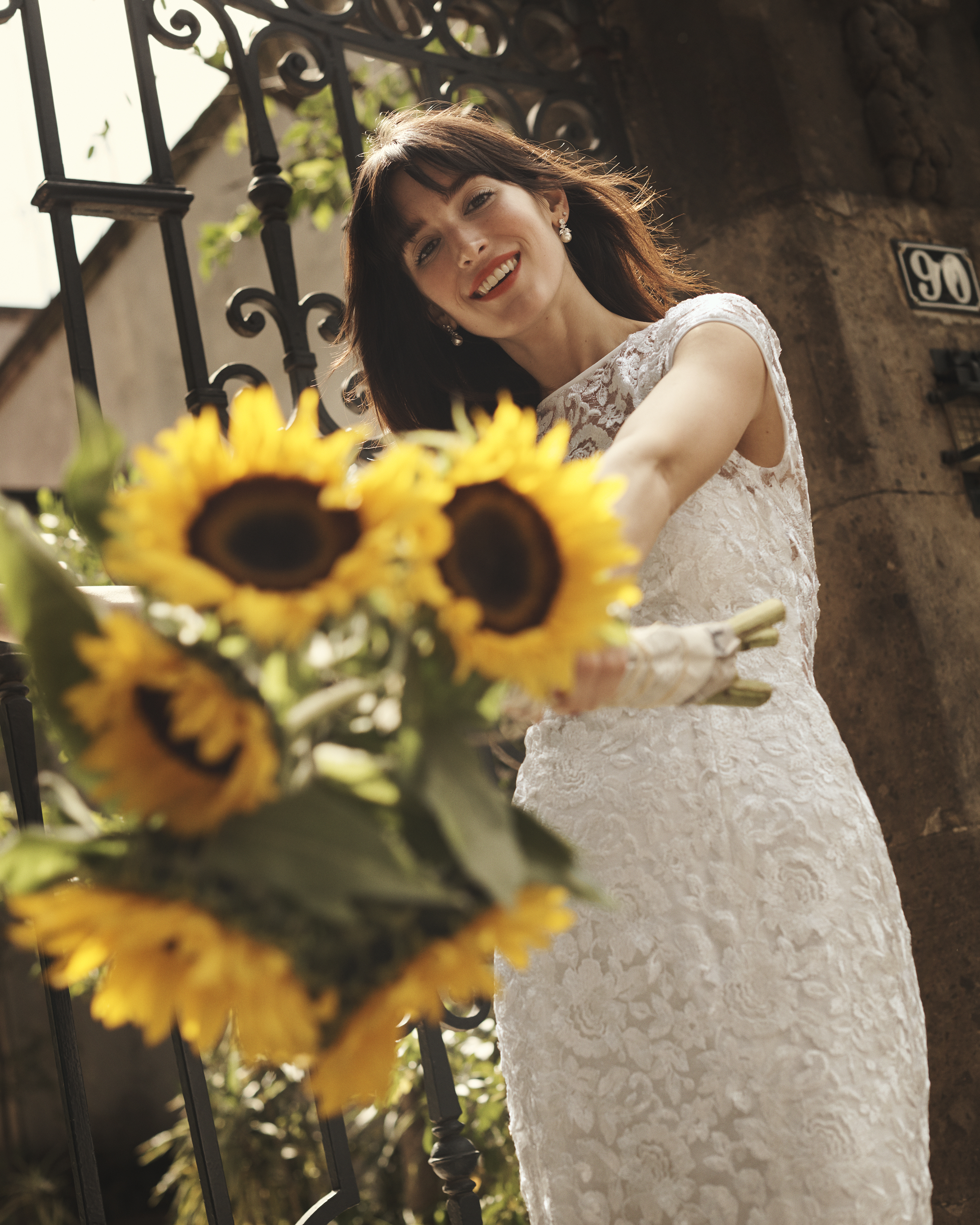 Smiling bride in long lace dress holding sunflowers towards camera.