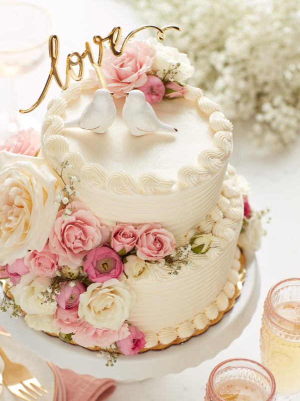 White two-tier wedding cake with pink and white flowers and a love script with two kissing birds cake topper