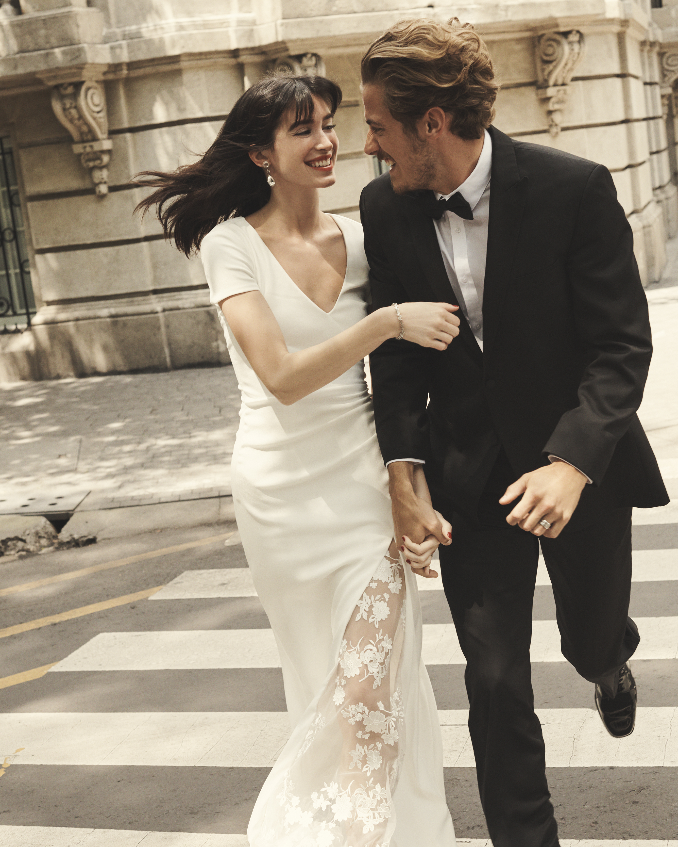 Smiling bride in white dress and groom in tux running hand in hand in street