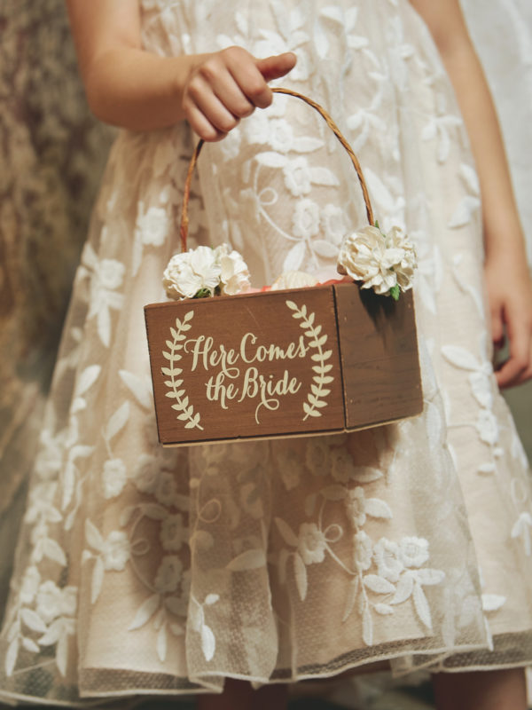 Flower girl in a lace dress holding a wooden basket with 'Here Comes the Bride' written on it