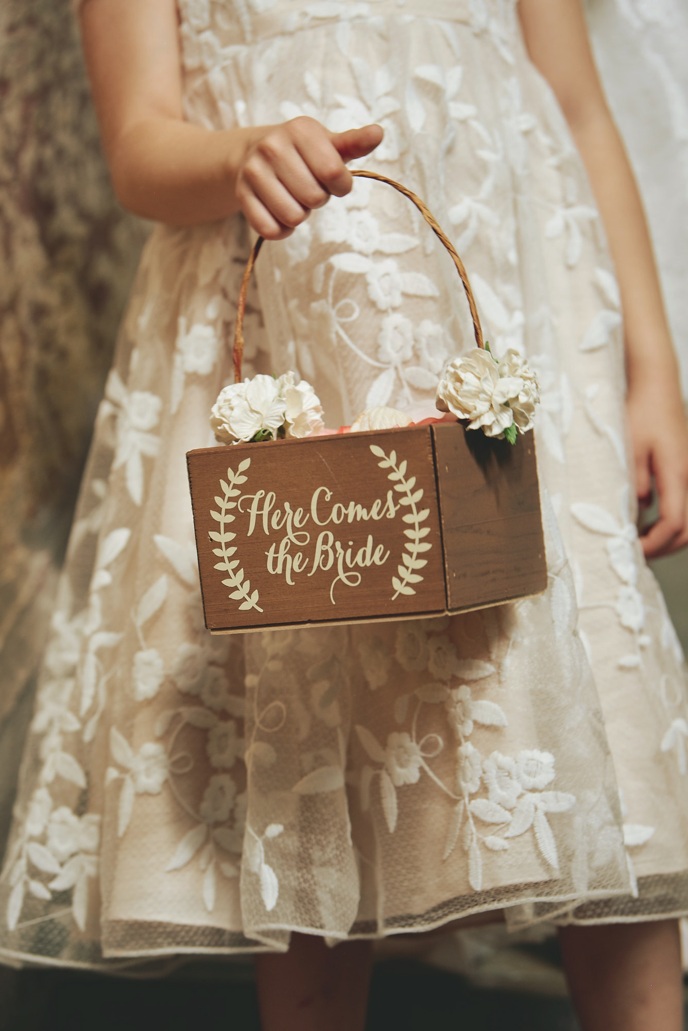 Flower girl in a lace dress holding a wooden basket with 'Here Comes the Bride' written on it