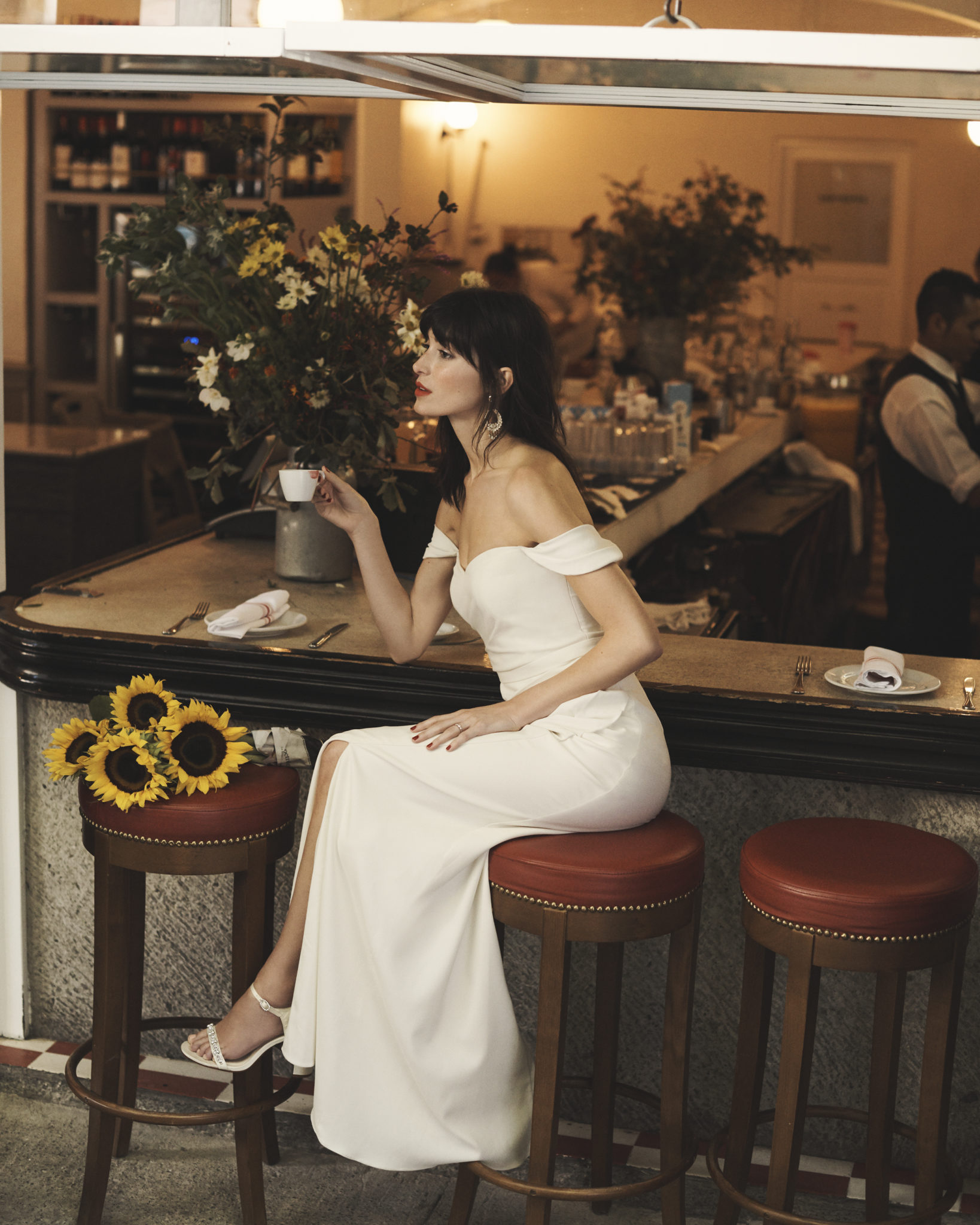 Bride sitting on stool in cafe wearing a draped off-the-should sheath wedding dress