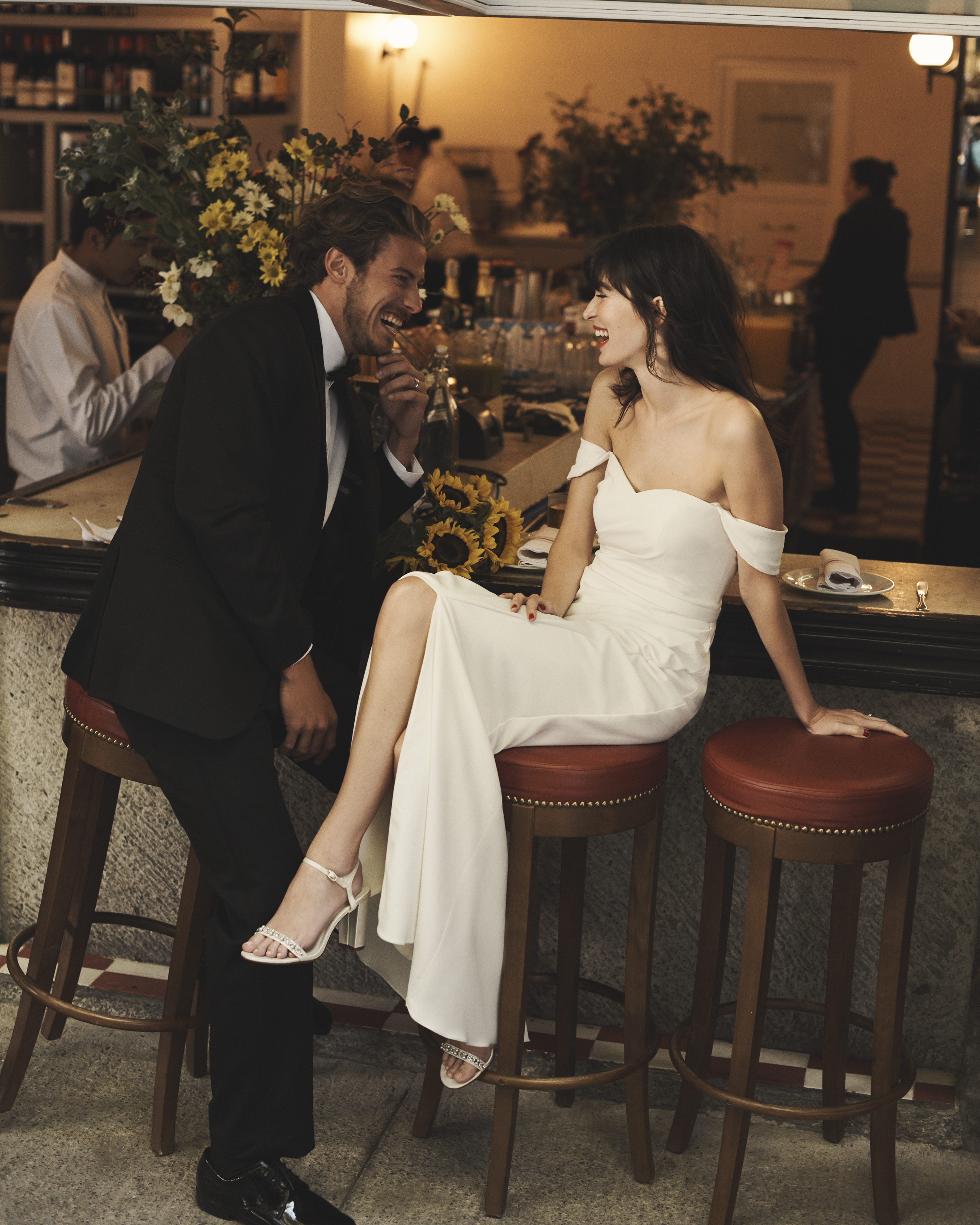 Groom in tux and bride in off-the-shoulder dress smiling in cafe