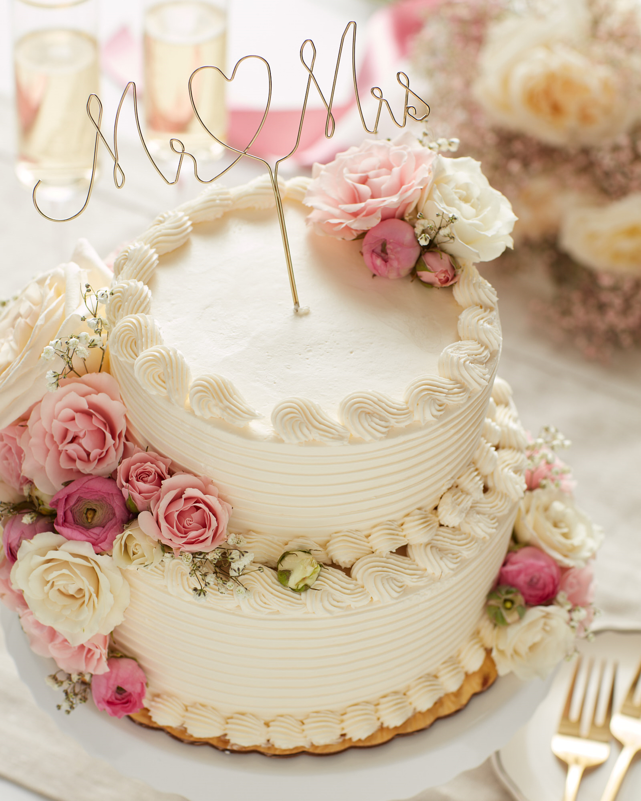 White two-tier wedding cake with pink and white flowers and a Mr and Mrs wire script with heart cake topper