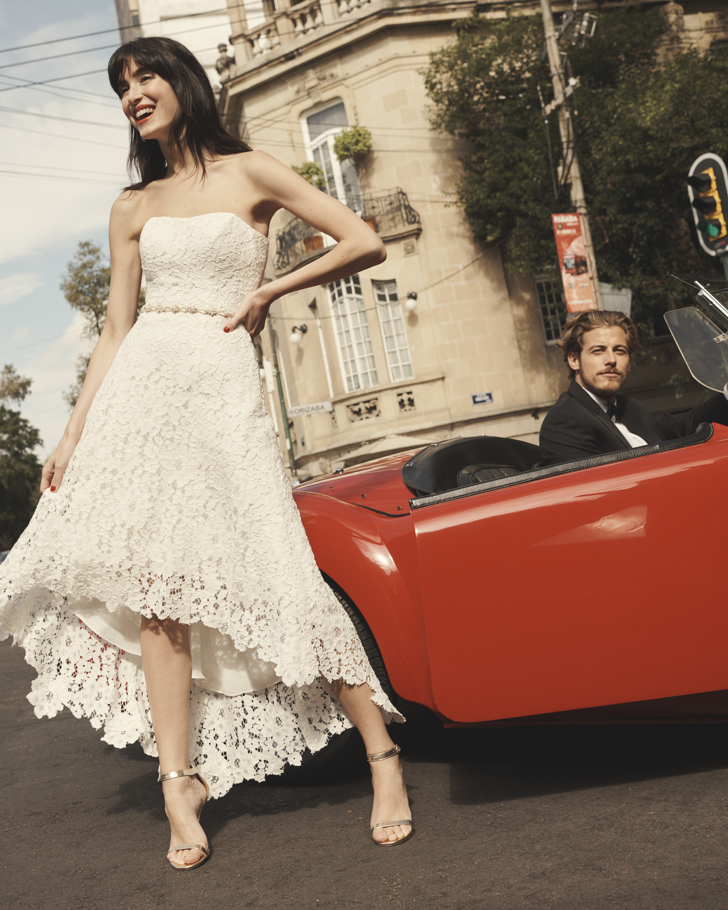 Smiling bride in strapless high-low lace dress on street in front of red convertible.