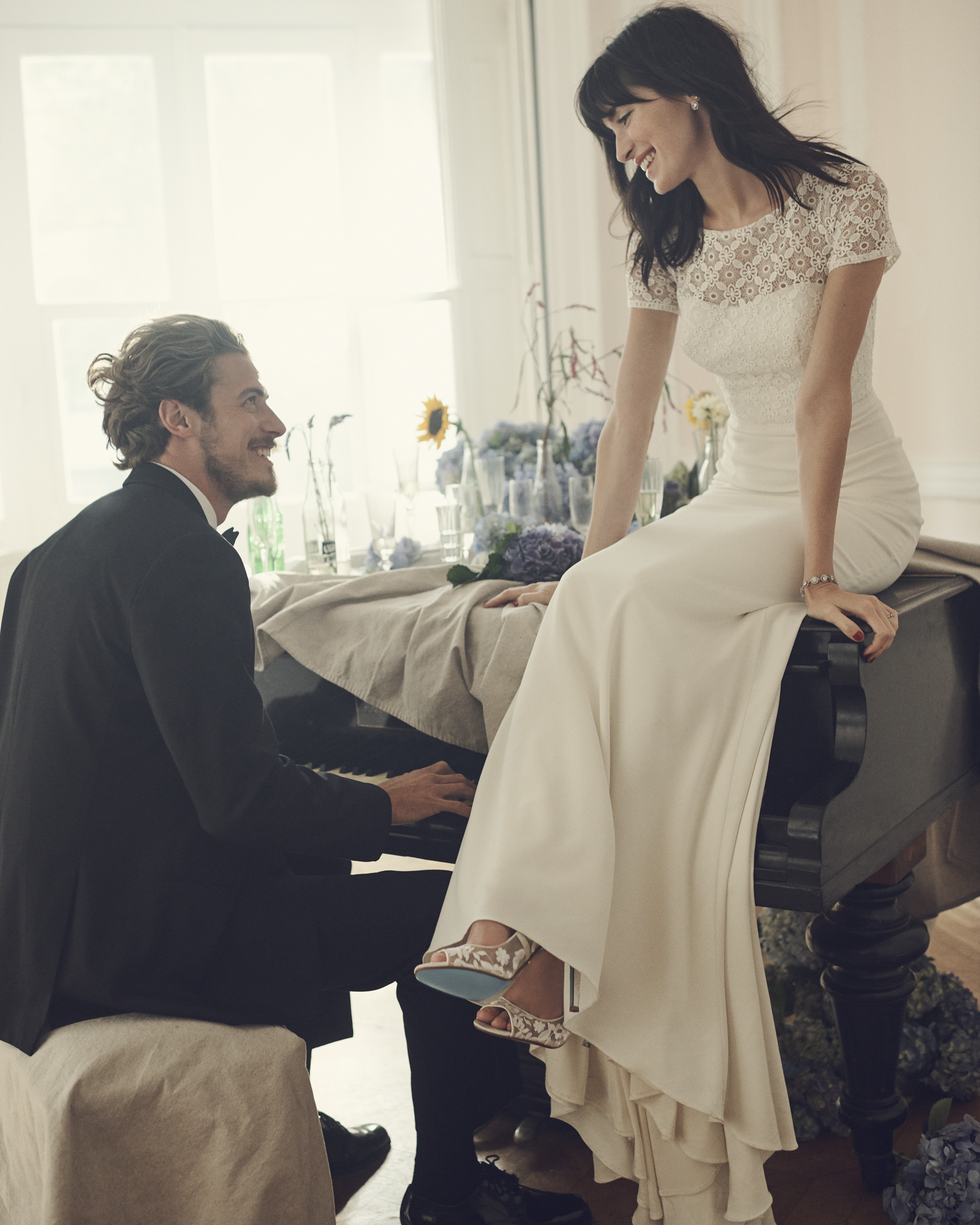 Bride in short sleeve wedding dress sitting on piano smiling at groom in tux.