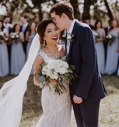 Groom kissing bride on the cheek with bridal party in background