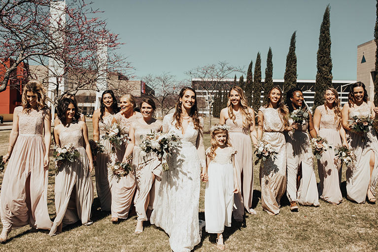 Bride walking with flower girl and 11 bridesmaids in beige and gold dresses.