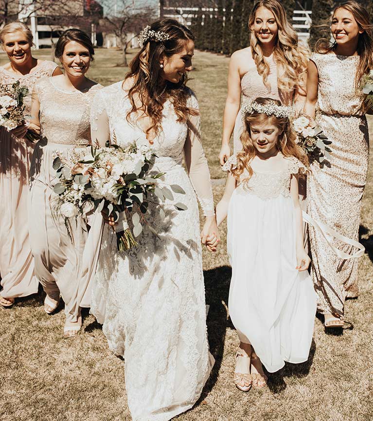 Bride holding hands with flower girl with bridesmaids in beige and gold dresses behind them