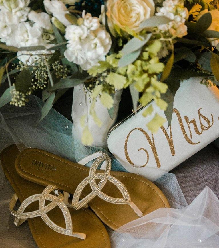 Bridal bouquet, ivory clutch with "Mrs" in gold glitter, and gold and crystal sandals on top of a crystal adorned veil. 