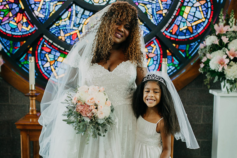 Bride with daughter in a white dress, tiara and short veil in front of a stained glass window