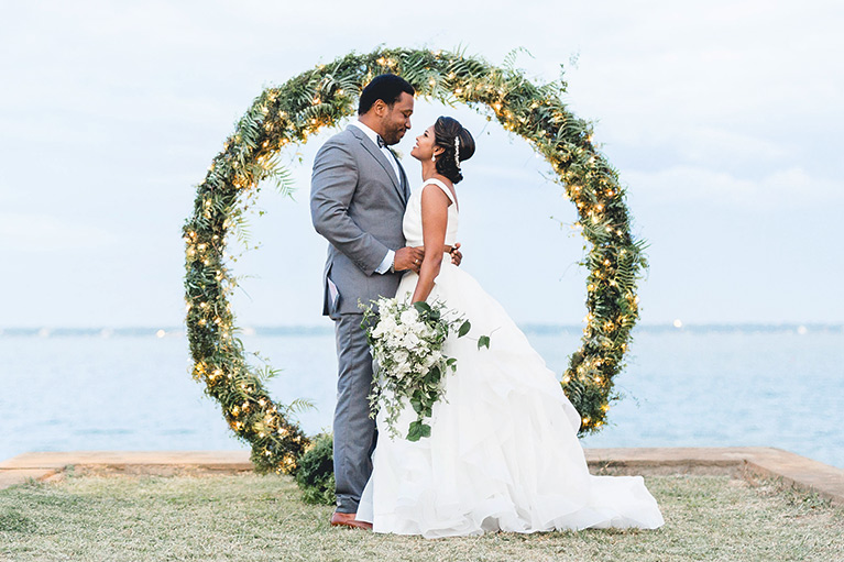 Bride and groom embracing behind a circle of greenery laced with twinkling lights and overlooking the sea