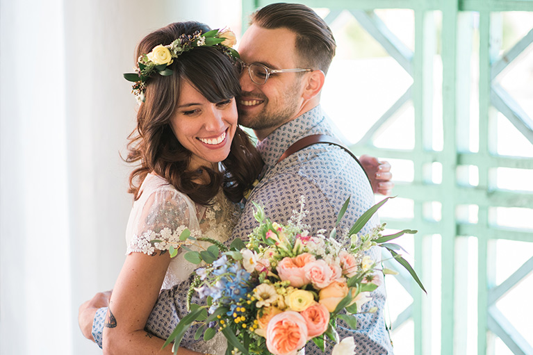 Bride and groom embracing. Bride wears a flower crown and holds a bouquet.