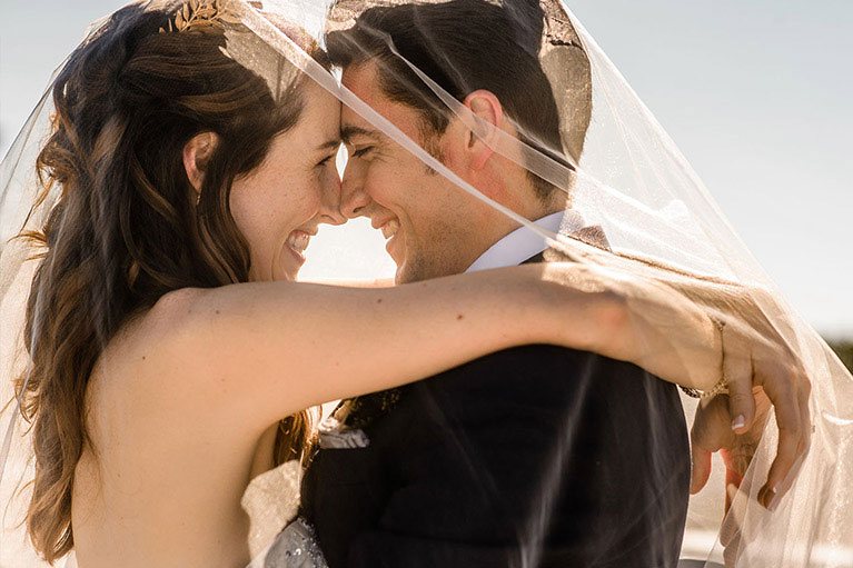 Close up portrait of bride and groom facing each other, foreheads touching underneath veil