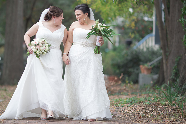 Two brides holding hands and their bridal bouquets while walking through a park