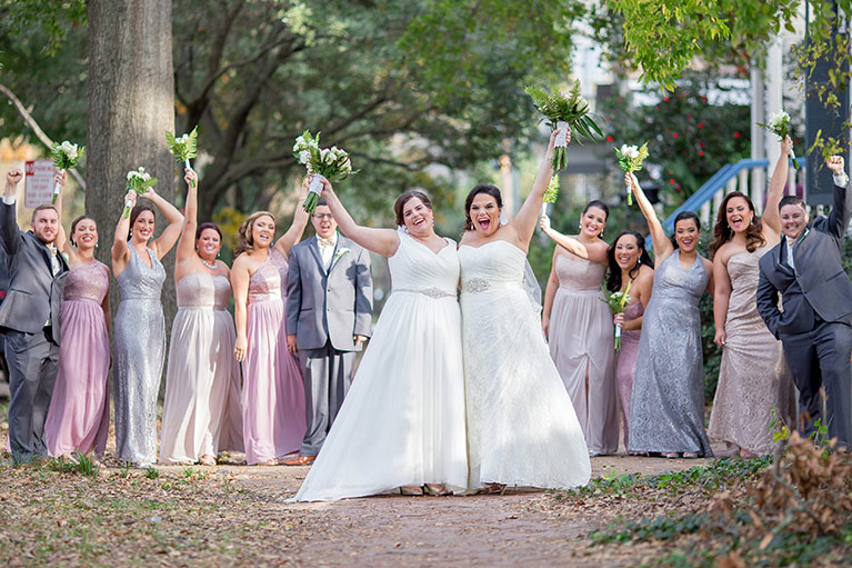 Two brides holding their bouquets in the air with their bridesmaids in light purple dresses and groomsmen in gray suits behind them