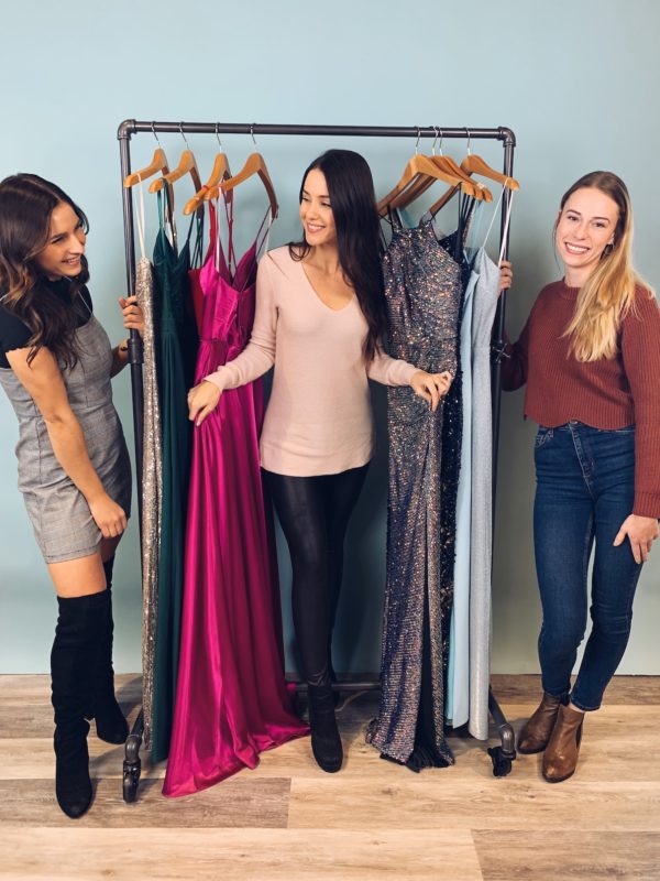 Girls smiling in front of prom dresses on a rack