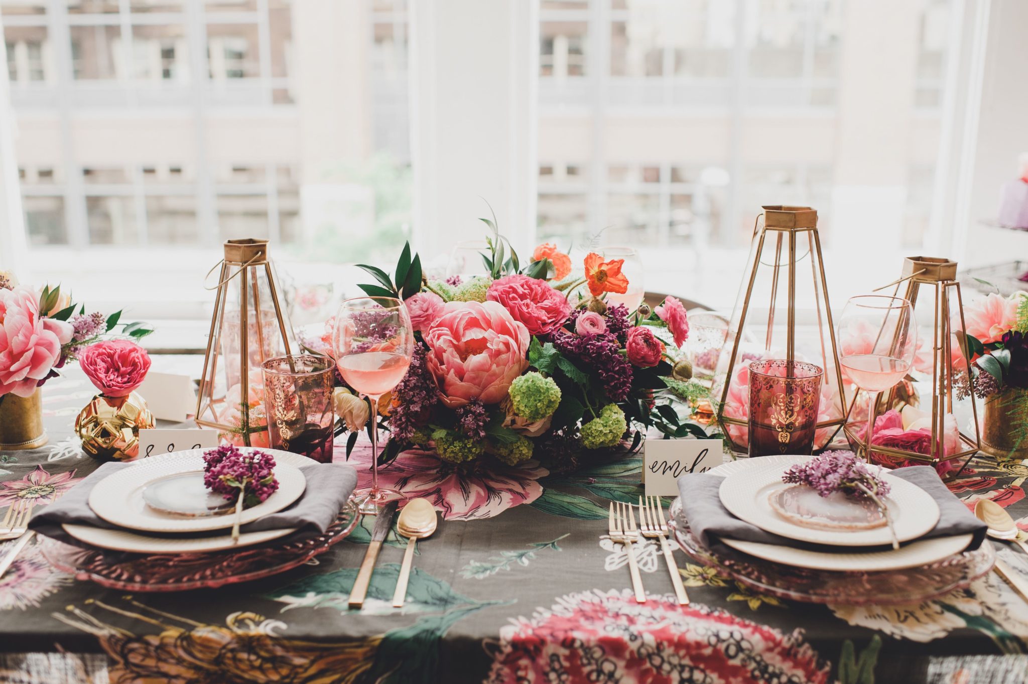 Table set with blue and pink tablecloth, white plates, gold flatware, gold candlesticks and pink flowers.