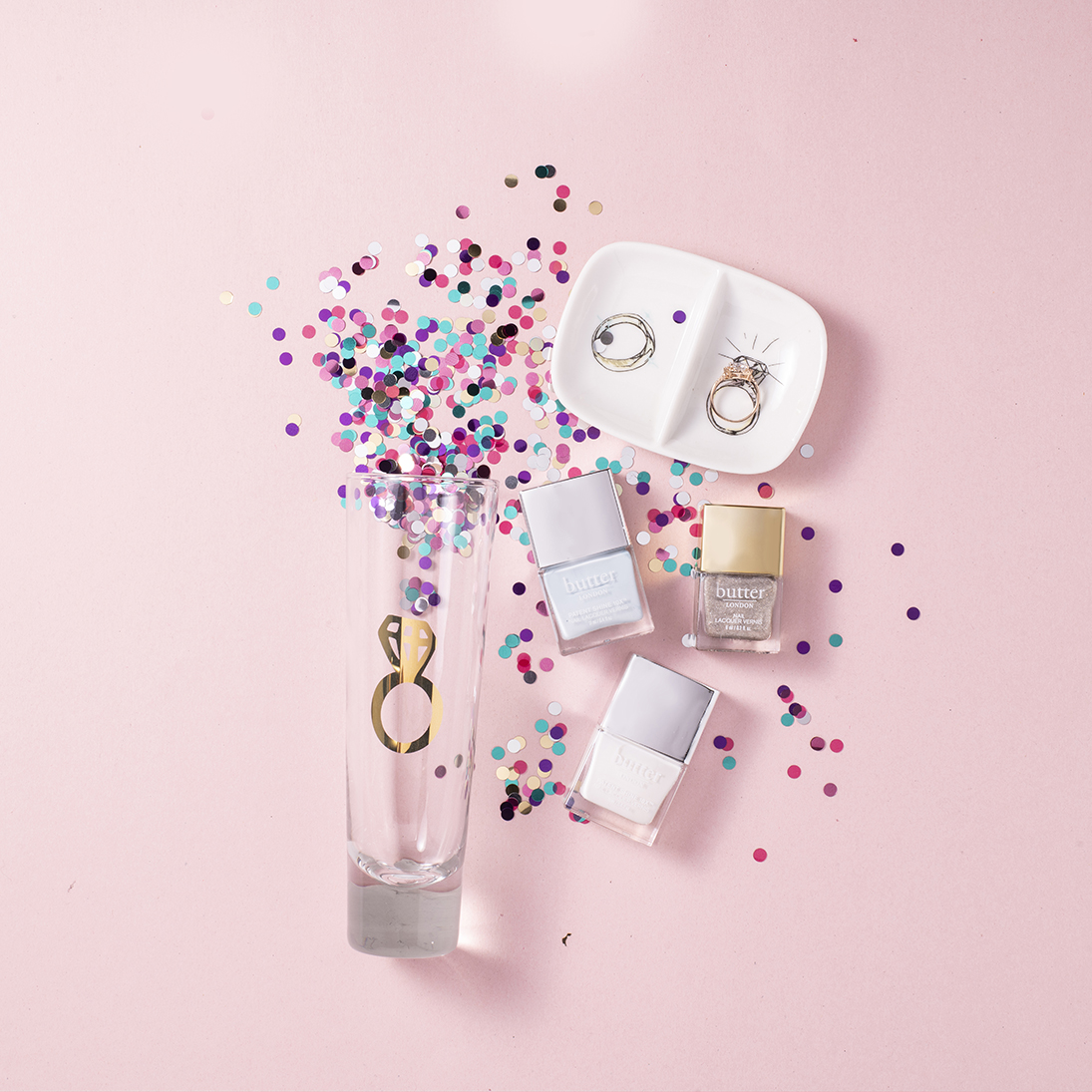 A champagne glass with confetti, a ring dish and nail polish bottles on a pink backdrop.