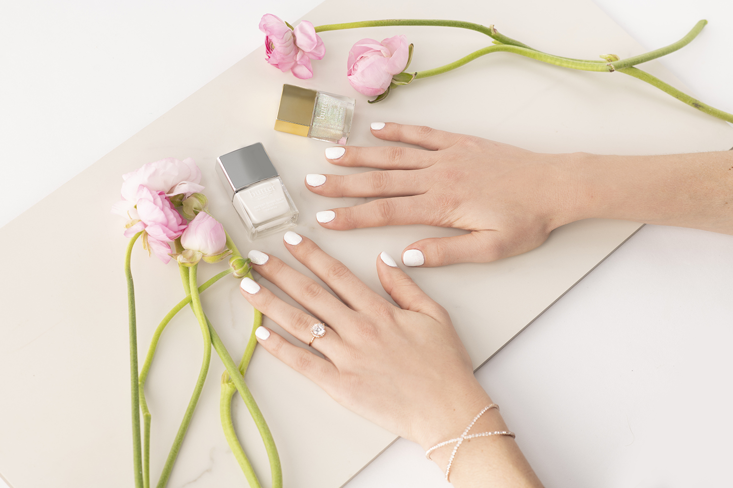 White and glitter nail art on marble with nail polish bottles and pink flowers.