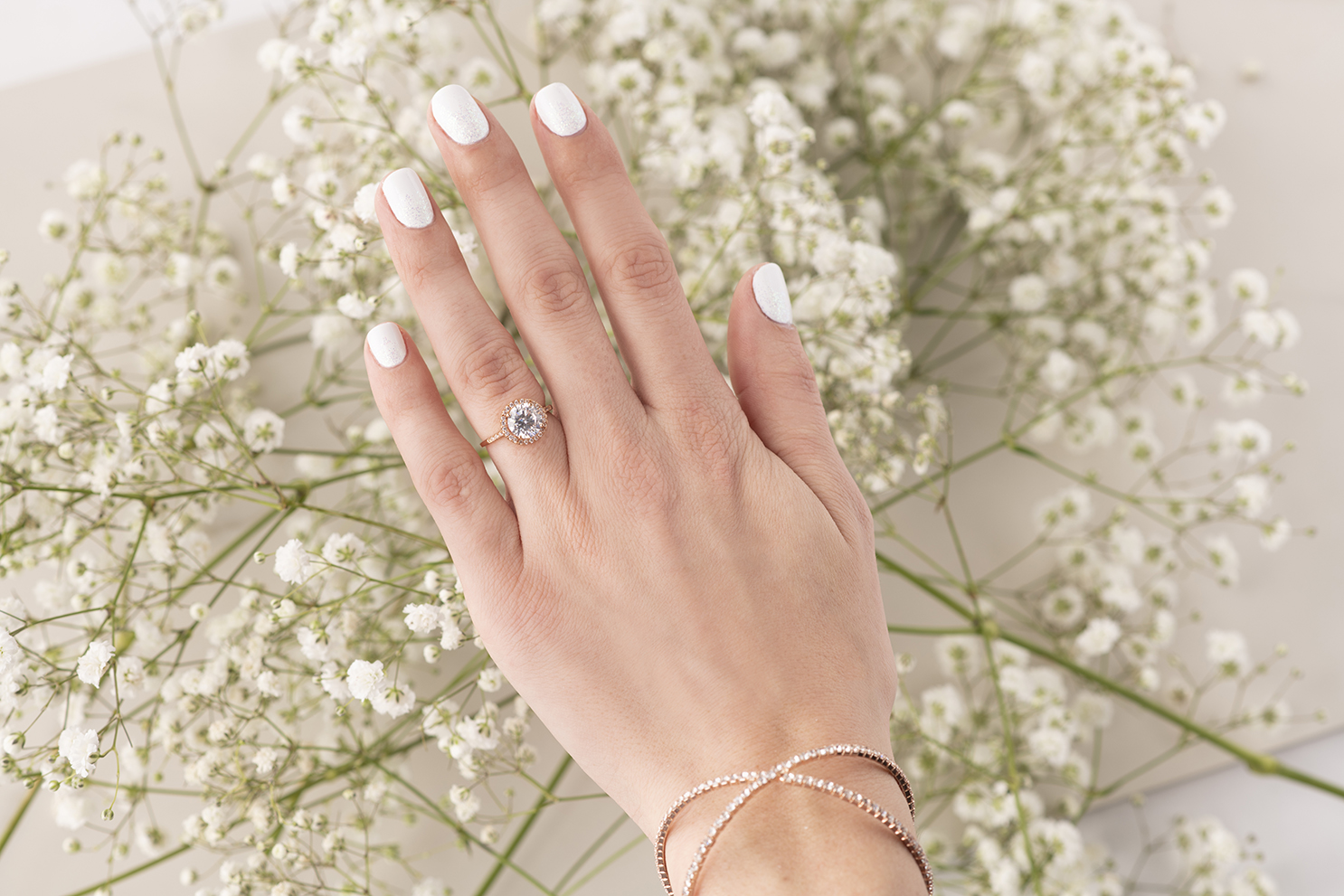 A woman's hand in front of white flowers with a white and glitter manicure.